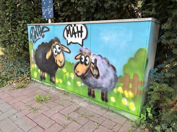 Ein Stromkasten, der mit Grafitti bemalt wurde mit einem schwarzen und einem weißen Schaf, die beide eine Sprechblase mit dem Wort MÄH haben.