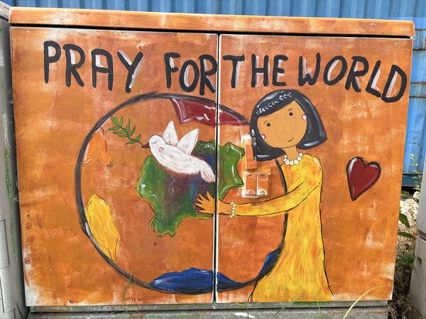 Ein Stromkasten, der mit Grafitti bemalt wurde und der eine Frau abbildet, die die Welt umarmt mit dem Schriftzug "Pray for the World"