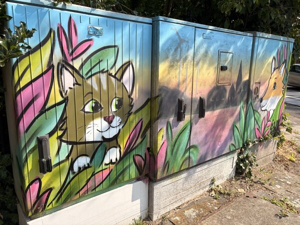 Drei Stromkästen, die mit Grafitti bemalt wurden und in der Mitte die Skyline von Schüttorf zeigen. Rechts ist ein Fuchs und links eine Katze abgebildet.