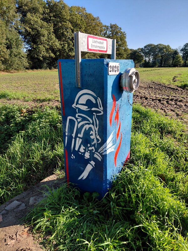 Ein Betonquader auf einem grünen Feld. Der Quader hat einen blauen Untergrund. Darauf steht ein Feuerwehrmann und löscht einen Brand.