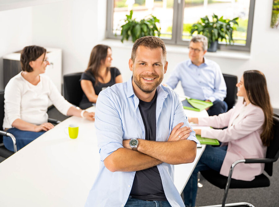 Mann steht am Tisch im Konferenzraum im Hindergrund sitzen vier Kollegen