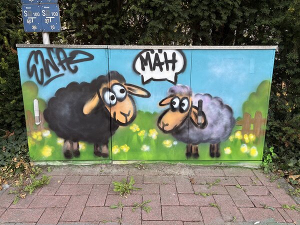Ein Stromkasten, der mit Grafitti bemalt wurde mit einem schwarzen und einem weißen Schaf, die beide eine Sprechblase mit dem Wort MÄH haben.