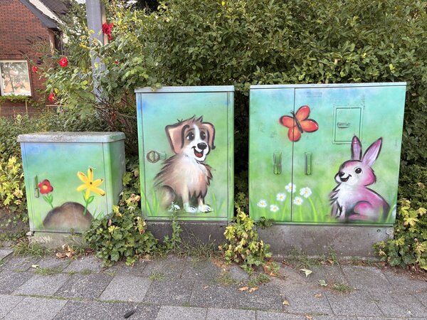 Drei Stromkästen, die mit Grafitti bemalt wurden und worauf ein Hund und ein Hase mit grüner Landschaft abgebildet sind.