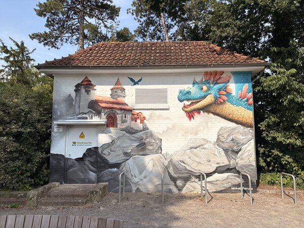 Eine großes Trafohaus, das mit Grafitti bemalt wurde und eine Burg und einen Drachen abbildet.