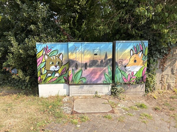 Drei Stromkästen, die mit Grafitti bemalt wurden und in der Mitte die Skyline von Schüttorf zeigen. Rechts ist ein Fuchs und links eine Katze abgebildet.