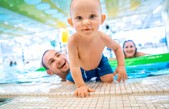 Baby krabbelt aus dem Schwimmbecken, Mutter und Vater lachen ihm zu